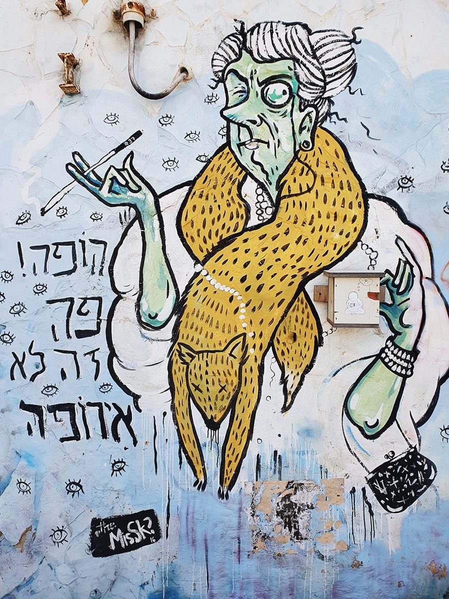 Street art mural of an elderly woman in a fur stole holding a cigarette holder, next to Hebrew graffiti reading הופה! פה זה לא אירופה ("Hopa! This isn't Europe here")