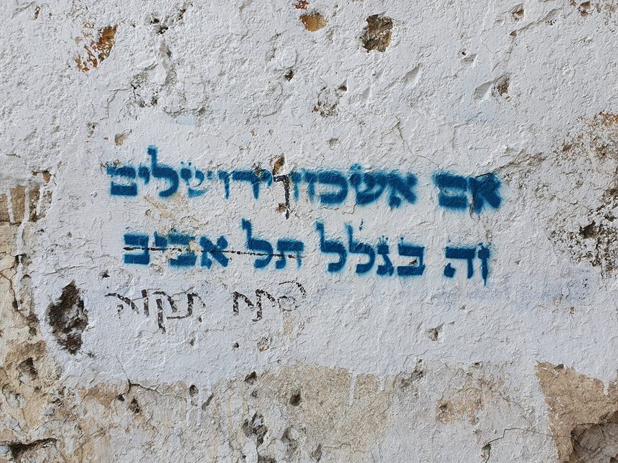 Blue stencil graffiti on a white wall reading אם אשכחך ירושלים זה בגלל תל אביב ("If I forget Jerusalem, it's because of Tel Aviv")