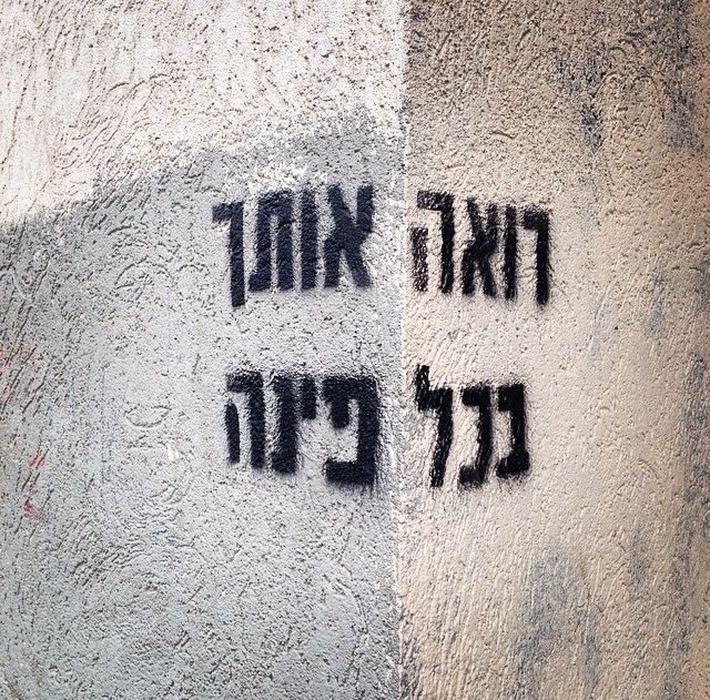 Black stencil graffiti on a split plaster wall reading רואה אותך בכל פינה ("Seeing you in every corner")